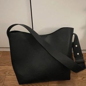 Svart axelväska - Säljer en stilren svart axelväska i syntetmaterial. Väskan har en enkel design med en bred axelrem och knappstängning. Perfekt för vardagsbruk och rymmer allt du behöver!