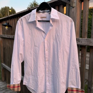 Vit skjorta från Burberry Brit - Säljer en stilren vit skjorta från Burberry Brit. Den har långa ärmar och klassiska knappar framtill. Skjortan är i bomull och har en broderad logga på bröstet. Manschetterna har ett snyggt rutigt mönster på insidan. Perfekt för både vardag och fest!