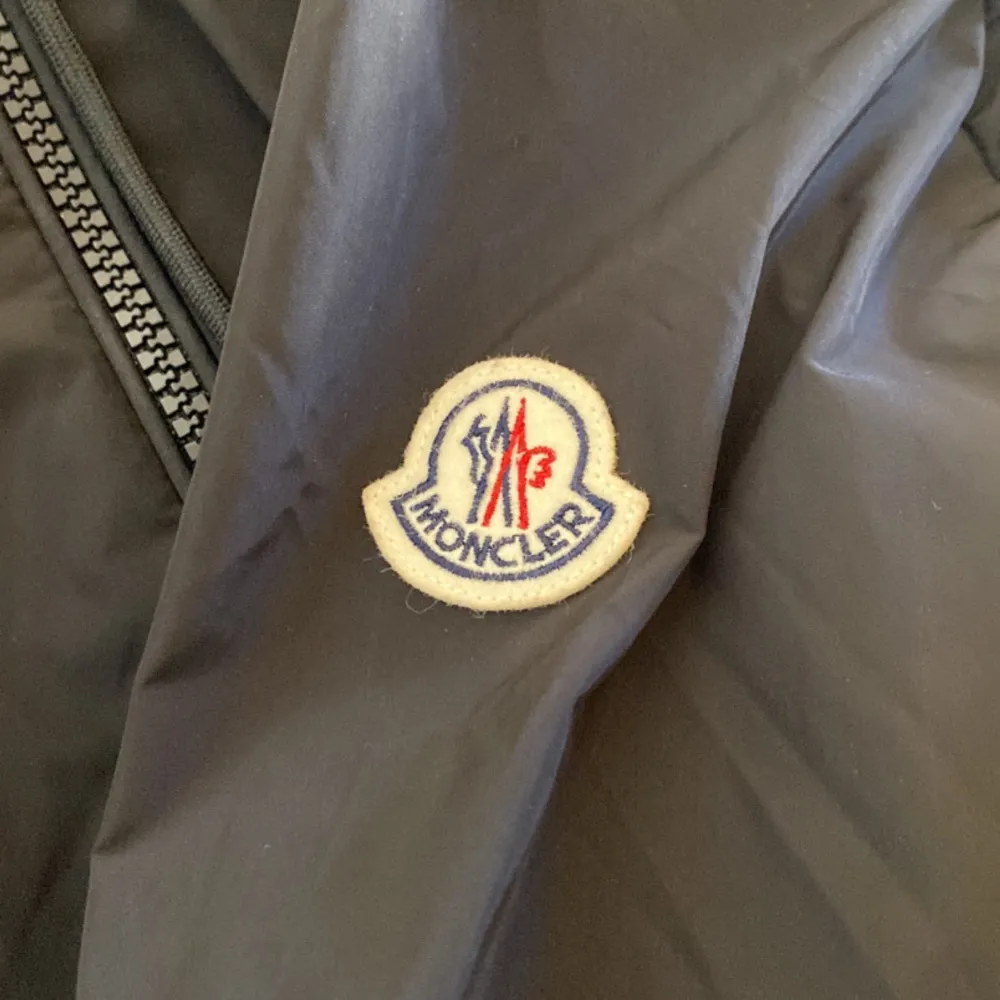 Säljer en stilren svart vindjacka från Moncler. Jackan har en dragkedja framtill och en huva med en snygg randig detalj. Perfekt för vår och höst när vädret är lite osäkert. Den är i bra skick och redo för nya äventyr!. Accessoarer.