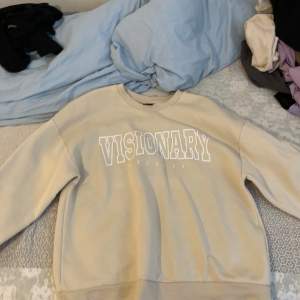 Säljer en snygg beige sweatshirt med texten 'VISIONARY' tryckt i vitt på framsidan. Tröjan har en klassisk rund hals och långa ärmar med ribbade muddar vid ärmslut och nederkant. Perfekt för en avslappnad stil!