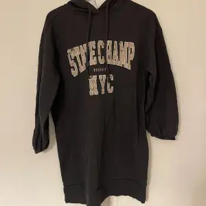 Säljer en svart hoodieklänning med texten 'Stone Champ NYC' på framsidan. Den har en klassisk design med långa ärmar och en bekväm passform. Perfekt för en avslappnad stil eller en dag på stan. 