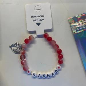 Handgjort armband med texten 'SAKNAD' - Ett superfint handgjort armband med röda och vita pärlor. Armbandet har bokstavspärlor som bildar ordet 'SAKNAD' och en liten hjärtpärla. Det finns också en silverfärgad berlock i form av en änglavinge. Perfekt som en personlig present eller för att bära med sig någon man saknar. 💖