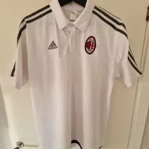 Säljer en snygg vit AC Milan fotbollströja från Adidas. Tröjan har korta ärmar och klassiska Adidas-ränder i svart på axlarna. Den har en broderad AC Milan-logga på bröstet och Adidas-loggan i grått. Perfekt för alla fotbollsfans!