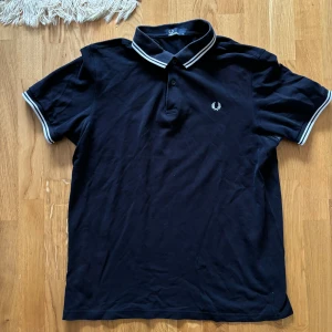 Svart pikétröja från Fred Perry - Säljer en snygg svart pikétröja från Fred Perry. Den har en klassisk design med vita ränder på kragen och ärmsluten. Tröjan är kortärmad och perfekt för både vardag och fest. Den är i bra skick och redo för en ny ägare!
