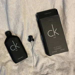 CK Be Eau de Toilette från Calvin Klein - Säljer en stilren och klassisk CK Be Eau de Toilette från Calvin Klein. Flaskan är mattsvart med en minimalistisk design och rymmer 100 ml. Doften är unisex och har en fräsch och aromatisk karaktär med toppnoter av lavendel, gröna noter, bergamott, mint, enbär och mandarin. Mellannoterna består av grönt gräs, persika, jasmin, fresia och magnolia. Perfekt för både vardag och speciella tillfällen!