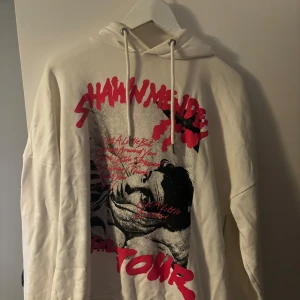 Vit hoodie från Shawn Mendes Tour - Säljer en supercool vit hoodie från Shawn Mendes Tour. Den har en stor, iögonfallande röd text och bild på framsidan och baksidan, vilket gör den perfekt för alla fans. Hoodien är långärmad och har en klassisk passform med en bekväm huva och dragsko. Perfekt för höst och vinter när du vill hålla dig varm och samtidigt visa din kärlek för Shawn Mendes!
