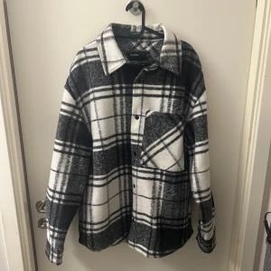 Svartvit rutig overshirt från Bershka - Snygg och trendig svartvit rutig overshirt från Bershka. Perfekt för lager-på-lager-stil med knappar framtill och en bröstficka. Den har en avslappnad passform och långa ärmar.