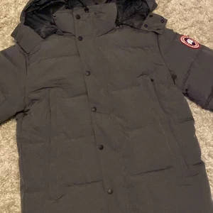 Svart dunjacka från Canada Goose - Säljer en svart dunjacka från Canada Goose med huva och knäppning framtill. Jackan har en klassisk design med varumärkets logga på ärmen. Perfekt för kalla vinterdagar.