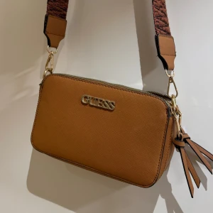 Axelväska från Guess - Snygg brun axelväska från Guess med justerbar axelrem i ett mönstrat tyg. Väskan har en dragkedja och en dekorativ tofsdetalj. Perfekt för att bära dina nödvändigheter med stil. (Väskan är inte äkta)💕