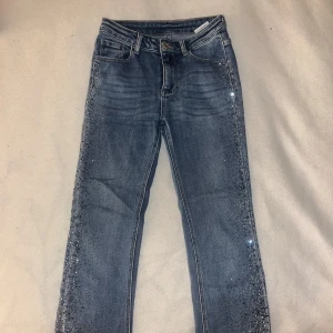 Blå jeans med paljettdetaljer - Snygga blå jeans med glittrande paljetter längs sidorna. Perfekt för att ge din outfit lite extra bling. Klassisk femficksdesign och knappgylf.