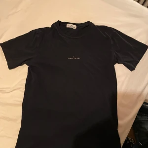 Mörkblå t-shirt från Stone Island - Snygg mörkblå t-shirt från Stone Island. Den har en diskret logga på framsidan och ett större tryck på baksidan. Perfekt för en stilren look.