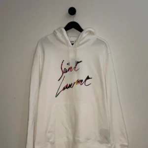 Saint Laurent Hoodie - Snygg vit hoodie från Saint Laurent med färgglatt tryck på bröstet. Den har en klassisk passform med långa ärmar och en justerbar huva. Perfekt för en stilren och avslappnad look. Bara testad fåtals gånger och kvittot finns, hör gärna av er vid flera frågor eller funderingar! 