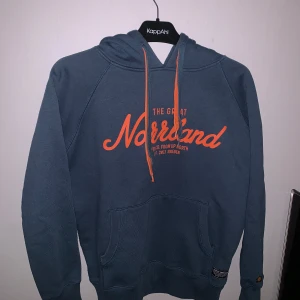 Blå hoodie från The Great Norrland - Säljer en blå hoodie från The Great Norrland med orange text på bröstet. Den har en stor ficka framtill och justerbar huva med orange snören. Perfekt för kyliga dagar!