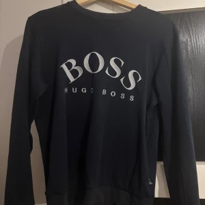 Svart tröja från Hugo Boss - Snygg svart tröja från Hugo Boss med stort vitt logotryck på framsidan. Tröjan har långa ärmar och en klassisk rund halsringning. Perfekt för en stilren look.