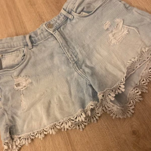 Ljusa jeansshorts med spetsdetaljer - Säljer ett par ljusa jeansshorts med snygga slitningar och spetsdetaljer längs kanten. Perfekta för sommardagar! De har en klassisk femficksdesign och knappgylf. Plagget är i bra skick priset kan diskuteras