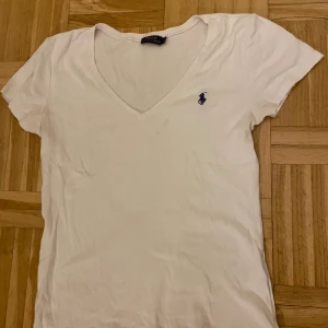 Vit t-shirt från Ralph Lauren - Säljer en klassisk vit t-shirt från Ralph Lauren med v-ringning och korta ärmar. Den har den ikoniska loggan broderad i blått på bröstet. Perfekt för en stilren och avslappnad look.