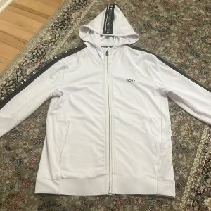 Vit huvtröja från Boss - Säljer en stilren vit huvtröja från Boss med svarta detaljer längs ärmarna och huvan. Tröjan har dragkedja framtill och långa ärmar. Perfekt för en avslappnad look.