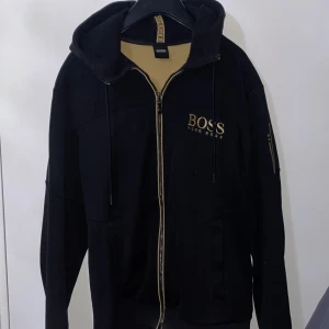 Svart hoodie från Boss - Snygg svart hoodie från Boss med dragkedja och guldfärgad logga på bröstet.  Mycket fin och finns inte längre att köpa i butik. nypris 1999