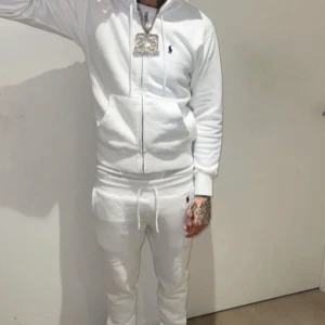 Vit tracksuit från Ralph Lauren - Säljer en stilren vit hoodie från Ralph Lauren med dragkedja och huva. Den har en liten broderad logotyp på bröstet och långa ärmar. Perfekt för en avslappnad look.