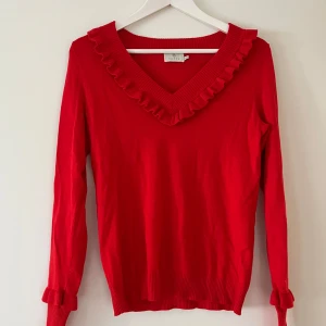 Röd tröja med volang från Kaffe - Säljer en snygg röd tröja från Kaffe med volangdetaljer vid v-ringningen och ärmsluten. Tröjan har långa ärmar och är perfekt för att liva upp garderoben. Passar bra till både jeans och kjol.