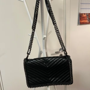 Svart quiltad axelväska med kedjedetaljer - Snygg svart axelväska i quiltat mönster med kedjedetaljer på remmen och kanterna. Perfekt för att ge en stilren touch till din outfit. Väskan har en praktisk storlek och är lätt att bära med sig.