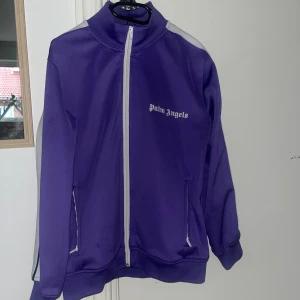 Lila träningsjacka från Palm Angels - Snygg lila träningsjacka från Palm Angels med vit dragkedja och vita ränder längs ärmarna. Jackan har en hög krage och två fickor framtill. Perfekt för en sportig look.