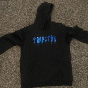 Svart hoodie från Trapstar - Säljer en svart hoodie från Trapstar med blått tryck på framsidan. Den har en klassisk design med huva och långa ärmar. Perfekt för en avslappnad stil.