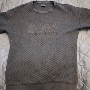 Svart tröja från Hugo Boss storlek S passar XS/S ny pris 1150  - Säljer en stilren svart quiltad tröja från Hugo Boss med broderad logga på bröstet. Tröjan har en rund halsringning och är perfekt för en avslappnad men snygg look.