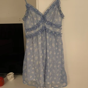 Sommarklänning med volangdetaljer - Söt blå klänning med blommigt mönster och volangdetaljer. Klänningen har smala axelband och en luftig passform, perfekt för sommaren. Sparsamt använd.