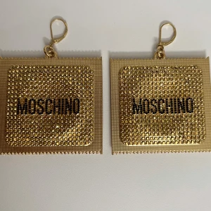 Guldiga örhängen från Moschino - Snygga och unika guldiga örhängen från Moschino. De är fyrkantiga med glittriga detaljer och Moschino-loggan i svart. Perfekta för att ge en lyxig touch till din outfit. Aldrig använda. 