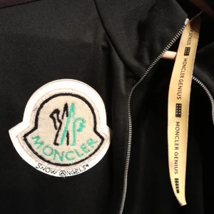 Svarta byxor från Moncler x Palm Angels - Snygga svarta byxor från Moncler x Palm Angels med lila sidopaneler och broderad logga. Byxorna har en elastisk midja för extra komfort och en avslappnad passform. Perfekta för en trendig och avslappnad stil. Jag själv har trivts i den då jag nu säljer den för hag växt ur den