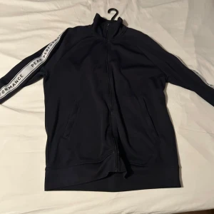 Svart hoodie från Peak Performance - Snygg svart hoodie från Peak Performance med dragkedja framtill. Den har långa ärmar med vita ränder och text längs ärmarna. Perfekt för en avslappnad stil.