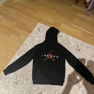 Svart hoodie från Nike - Säljer en svart hoodie från Nike med det ikoniska Jordan-trycket i rött och vitt på framsidan. Tröjan har en klassisk känguruficka och en bekväm huva. Perfekt för en sportig look!