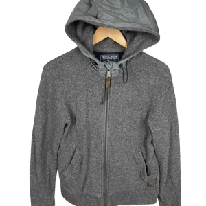 Grå jacka med huva från Woolrich - !INTRESSESKOLL! denna grå jacka från Woolrich med en praktisk huva och dragkedja framtill. Jackan har ribbade muddar och två fickor för extra komfort. Perfekt för kyligare dagar. 