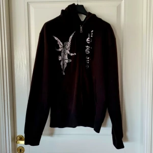 Svart hoodie från One of One - Säljer en svart hoodie från One of One med ett unikt tryck av en ängel på framsidan. Tröjan har en dragkedja vid halsen och långa ärmar. Perfekt för vintern 