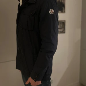 Moncler Jacka - Snygg svart jacka från Moncler med dragkedja och knappar. Jackan har två bröstfickor med lock och långa ärmar. Perfekt för en stilren look. Liten fläck vid nedre mitt av jackan + knappen längst ner saknas