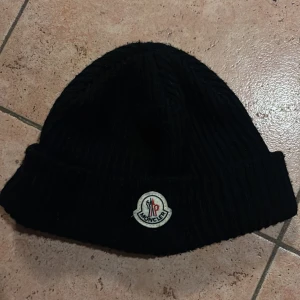 Svart stickad mössa från Moncler - Snygg svart stickad mössa från Moncler med broderad logga framtill. Perfekt för kyliga dagar och ger en stilren look. Passar både till vardags och mer uppklädda tillfällen.