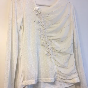 Vit långärmad blus med spetsdetaljer - Elegant vit blus med långa ärmar och vackra spetsdetaljer. Blusen har en unik design med en blomapplikation och subtila mönster längs framsidan. Perfekt för en stilfull look.