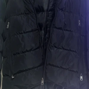 Moncler jakke  - Sort jakke med hætte og lynlåslukning. Jakken har Moncler logo på brystet og på ærmerne. Der er også et lille Moncler logo på venstre side af jakken. Jakken har to lommer foran og en lomme på venstre ærme. Der er en lille lomme på indersiden af jakken. Jakken har en snor i hætten, som kan strammes. Kun brugt en vinter