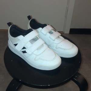 Vita sneakers med kardborreband från Adidas - Snygga vita sneakers från Adidas med praktiska kardborreband för enkel på- och avtagning. Skorna har en klassisk design med svarta detaljer och en bekväm passform. Har använt 2 gånger. Säljer för att dom är för små för mig💞
