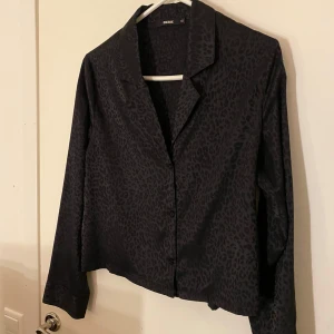 Svart leopardmönstrad skjorta från Bik Bok - Snygg svart skjorta från Bik Bok med leopardmönster. Skjortan har långa ärmar och knappar framtill, perfekt för en trendig look.
