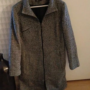 Grå kappa från XLNT Woman stl 44/46 - Snygg grå kappa från XLNT Woman med dragkedja framtill. Den har en klassisk krage och två fickor och en ficka på högra bröstet, alla med dragkedja. Perfekt för kyligare dagar.passar både till fest och vardags.