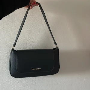 Svart axelväska från Valentino - Elegant svart axelväska från Valentino i skinn med en stilren design. Väskan har en praktisk dragkedja och en magnetisk knappstängning. Perfekt för att bära dina nödvändigheter med stil.