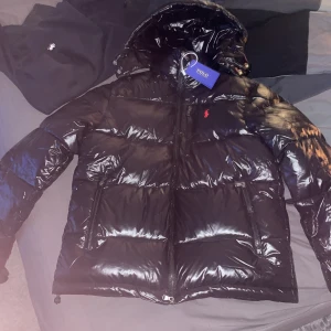 Svart dunjacka från Polo Ralph Lauren - Snygg svart dunjacka från Polo Ralph Lauren med glansig finish och huva. Jackan har dragkedja och två praktiska fickor framtill. Perfekt för kyliga dagar.