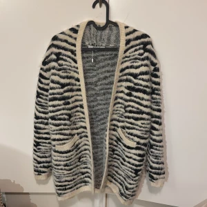 Zebramönstrad kofta från Easy Wear - Säljer en fluffig och mysig zebramönstrad kofta från Easy Wear. Koftan har en öppen design med två fickor framtill och långa ärmar. Perfekt för att hålla sig varm och stilren under kyliga dagar.