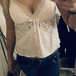 Rosa spetslinne med rosett - Säljer ett sött rosa linne med spetsdetaljer och en liten rosett framtill. Linnet har justerbara axelband och en luftig passform, perfekt för varma dagar.