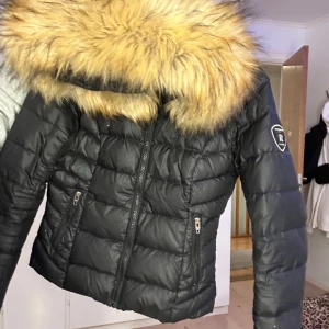 Svart dunjacka från Rockandblue - Snygg svart dunjacka från Rockandblue med en stor, fluffig pälskrage. Litet hål längst ner på ena armen men inget som syns. Köptes för ca 3000kr på rea och säljer för 400kr