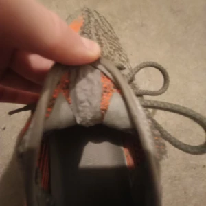 Adidas Yeezy Boost 350 V2 i grå och orange - Säljer ett par Adidas Yeezy Boost 350 V2 i grått med en iögonfallande orange rand och texten 'SPLY-350' på sidan. Skorna har en stickad ovandel och en ribbad sula för extra komfort. Perfekta för dig som vill ha en unik stil. Skorna är inte ekta.. använda sparsamt. Orginal pris 1800..