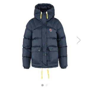 Mörkblå dunjacka från Fjällräven - Säljer en mörkblå dunjacka från Fjällräven med huva och gula detaljer. Jackan har en pufferdesign med knappar och dragkedja framtill samt praktiska fickor. Perfekt för kalla dagar.