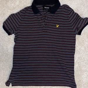 Randig pikétröja från Lyle & Scott - Snygg randig pikétröja från Lyle & Scott med klassisk krage och knappar. Tröjan har en broderad logga i gult på bröstet och korta ärmar. Perfekt för en avslappnad stil.
